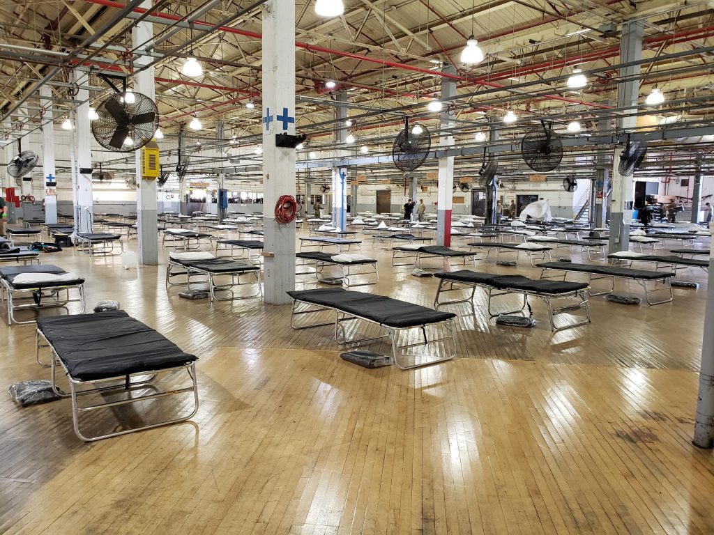 Inside of the alternative care site(ACS) with black foldable beds spread apart in a large room