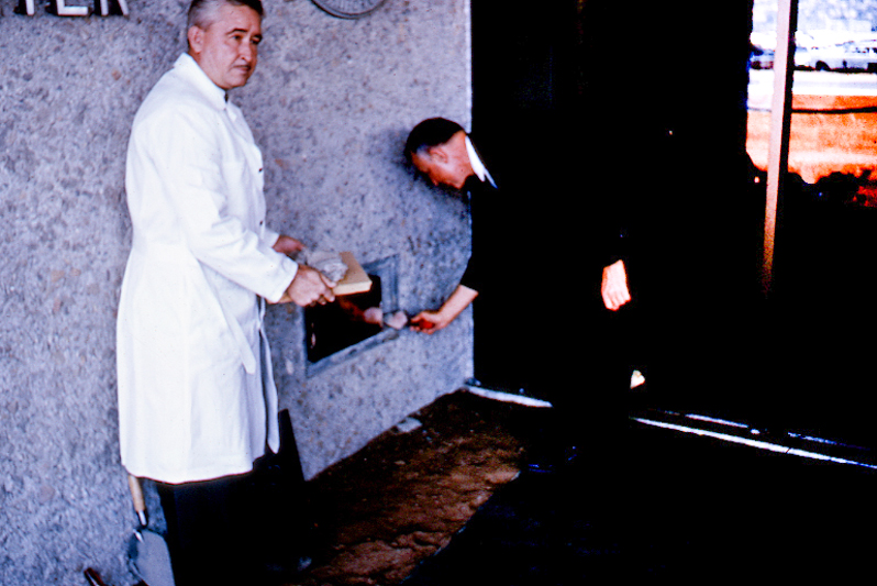 Two men stand near door to a prison, one of them is cementing something into the wall.