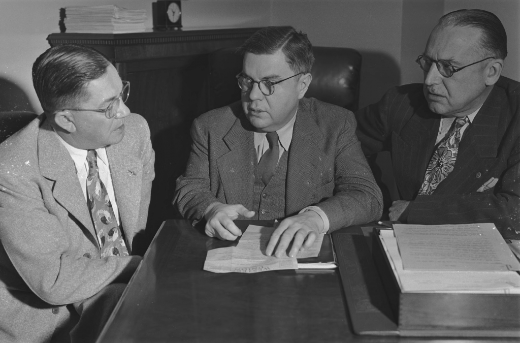 Three men sit at a table with papers around them. 