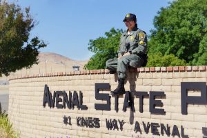 Woman in correctional uniform sits on a wall that says Avenal State Prison.
