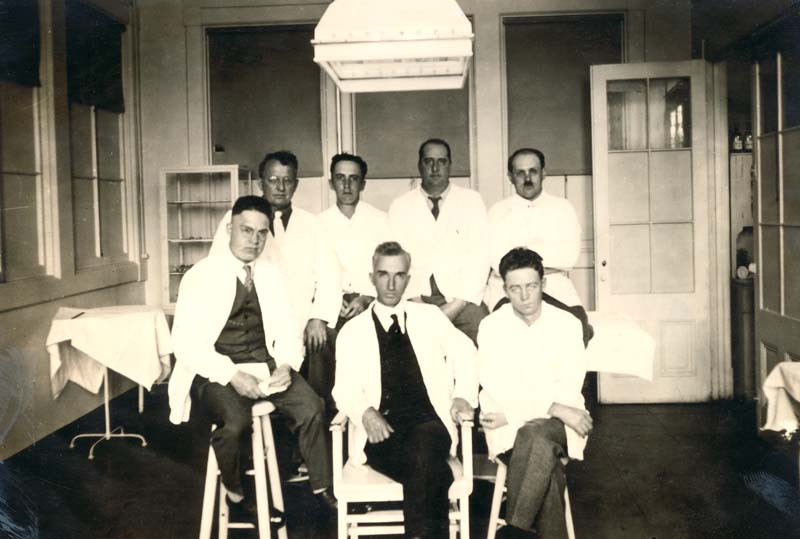 Seven men in lab coats pose for a photo in a prison doctor's office.