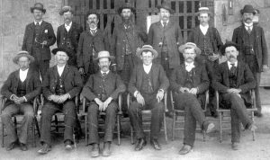 Men in hats, jackets and ties sit or stand in front of a prison gate.