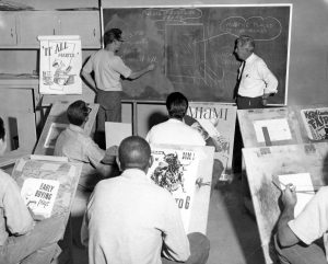 Two men at a chalkboard while others sit in desks and watch.