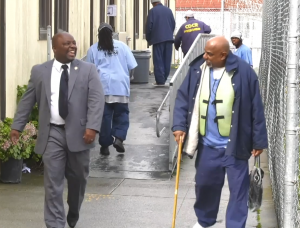 Smiling man in tie and jacket walks by another man with a cane.
