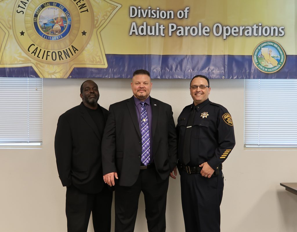 Three men stand in front of a Division of Adult Parole Operations banner.
