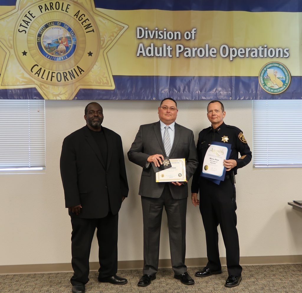 Three men stand in front of a banner that says Division of Adult Parole Operations and State Parole Agent California.