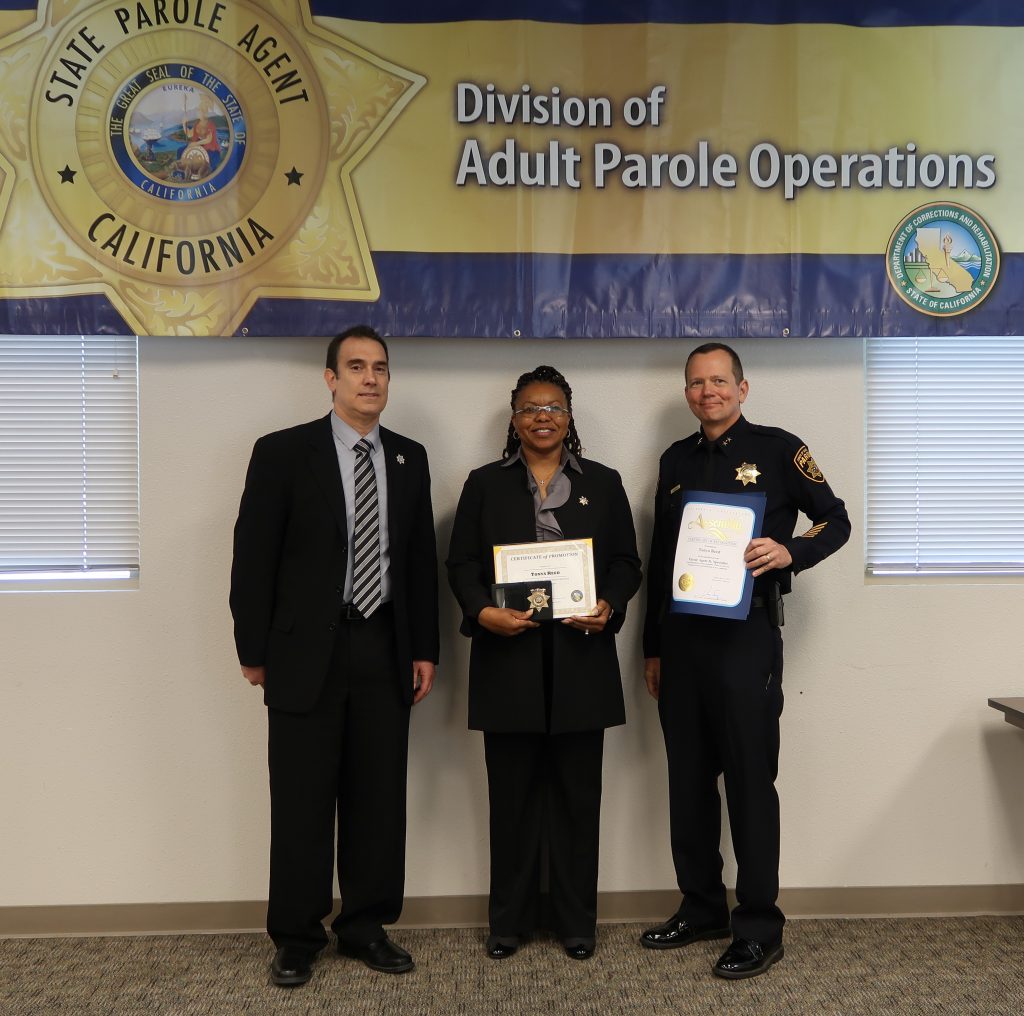 Two men and a woman stand in front of a banner that says Division of Adult Parole Operations and State Parole Agent California.