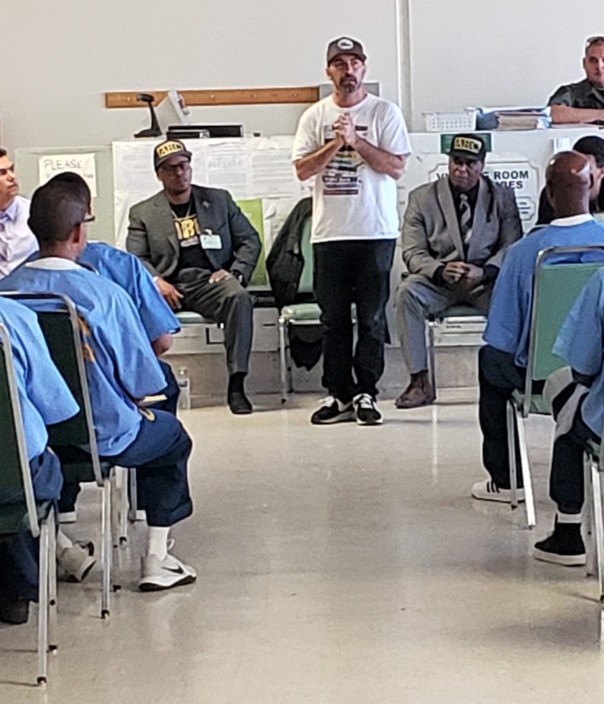 Man in tee-shirt speaks to men in inmate uniforms.