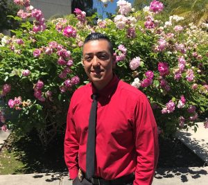 Man in red shirt and dark tie stands in front of rose bushes.