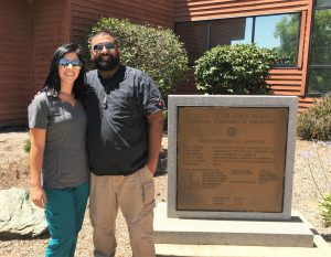 Woman in scrubs and man in regular clothing stand in front of the plaque for Mule Creek State Prison.
