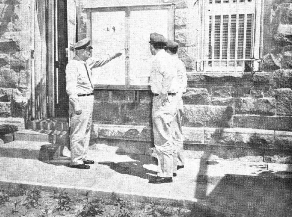 Three uniformed men stand in front of a brick wall with flyers on a board. One man points to them.