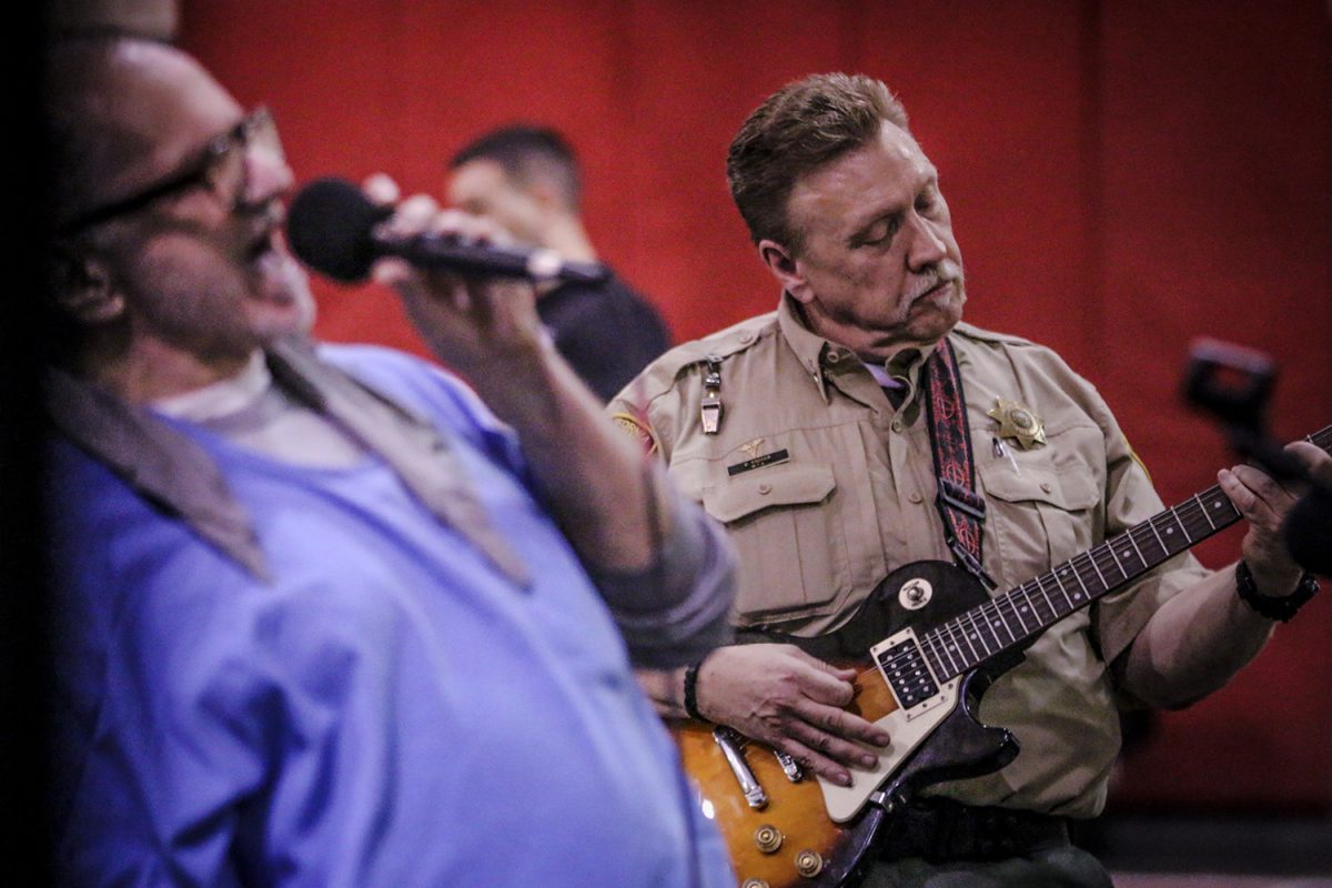 As music therapy, a man in blue shirt sings in microphone while correctional officer plays guitar.
