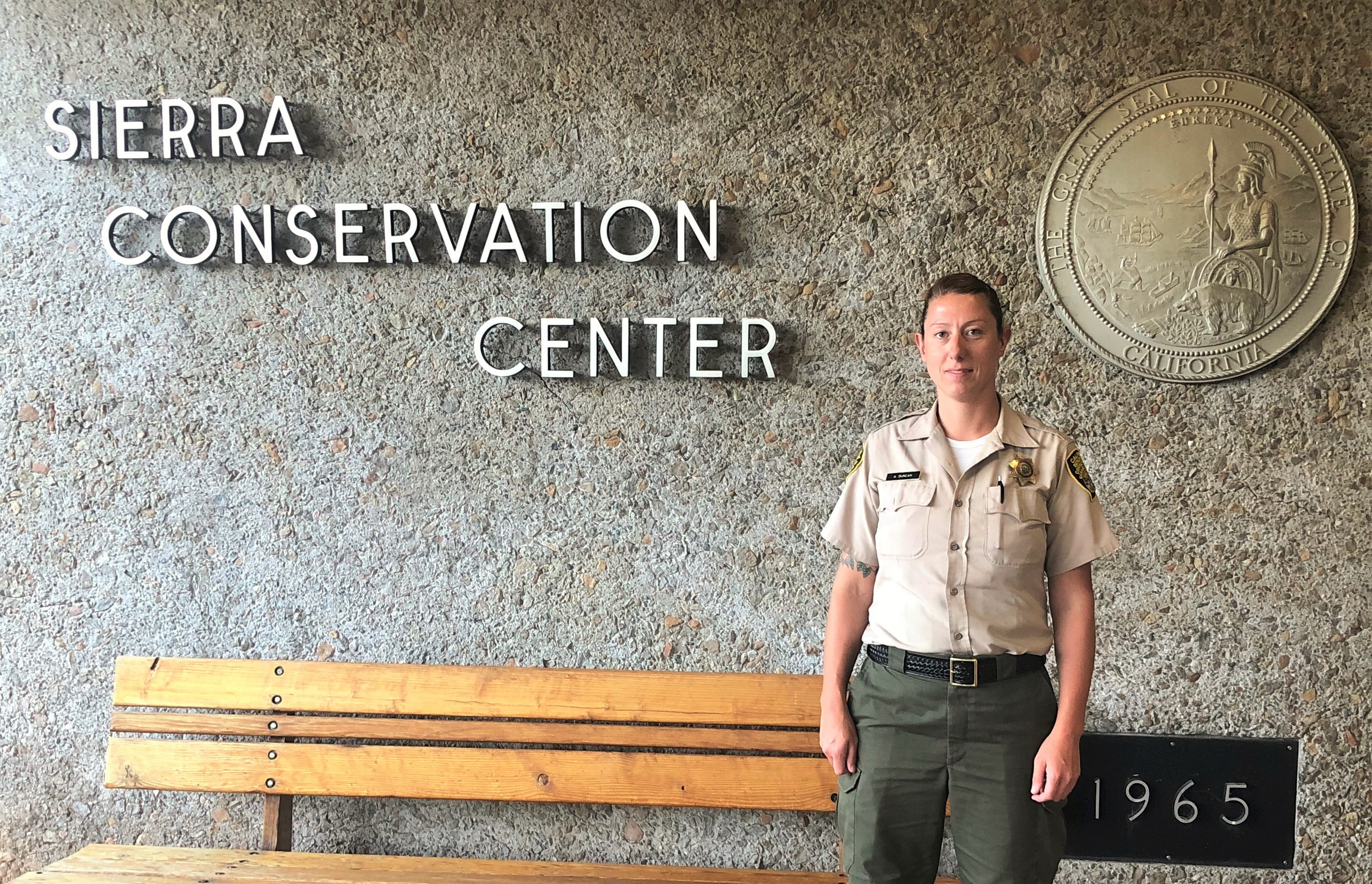 Woman in correctional officer uniform stands in front Sierra Conservation Center sign.