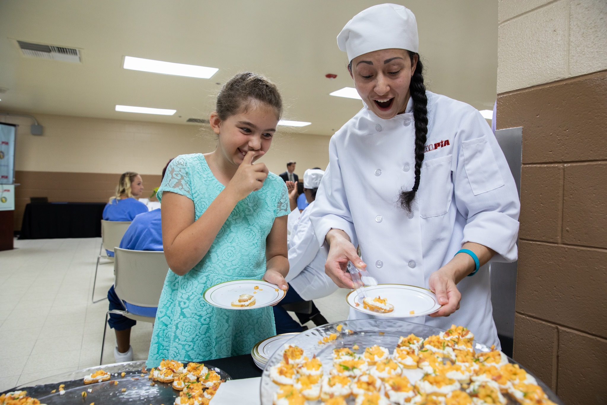 Woman in chef's clothing and young girl sample food.