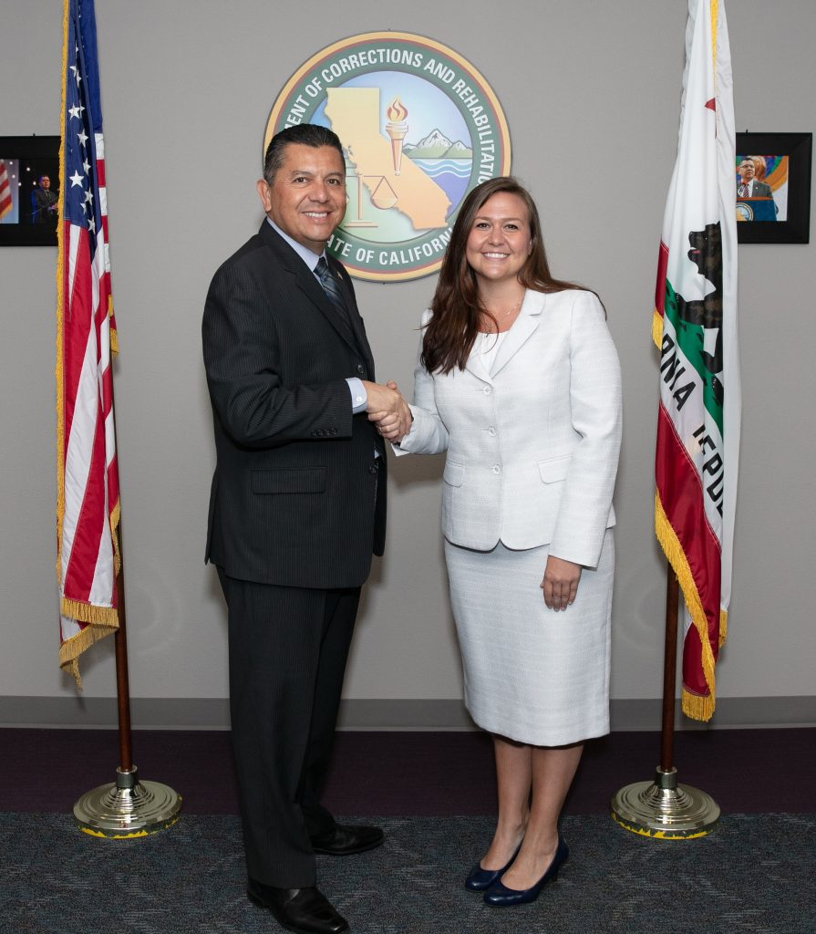 Man shakes hands with woman standing in front of CDCR logo and flags.