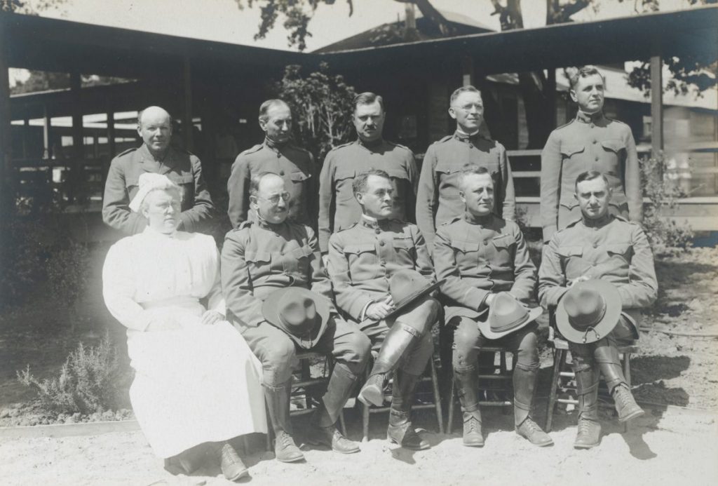 Men in uniform sit in front of a building with one nurse wearing white. 