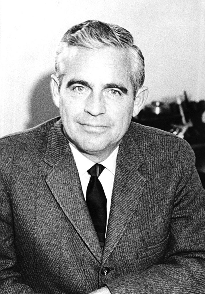 Black and white photo of man with graying hair, wearing a jacket and tie.
