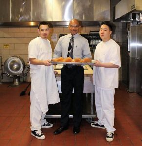 Man wearing tie holds a tray of rolls along with two young men in white clothing. They are standing in a kitchen.