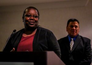 Woman speaks into microphone while a man stands behind her.