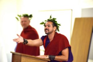 Men wear togas, while one gestures to the audience.