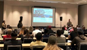 A roomful of sitting people watching a slide on a screen at the front of the room.
