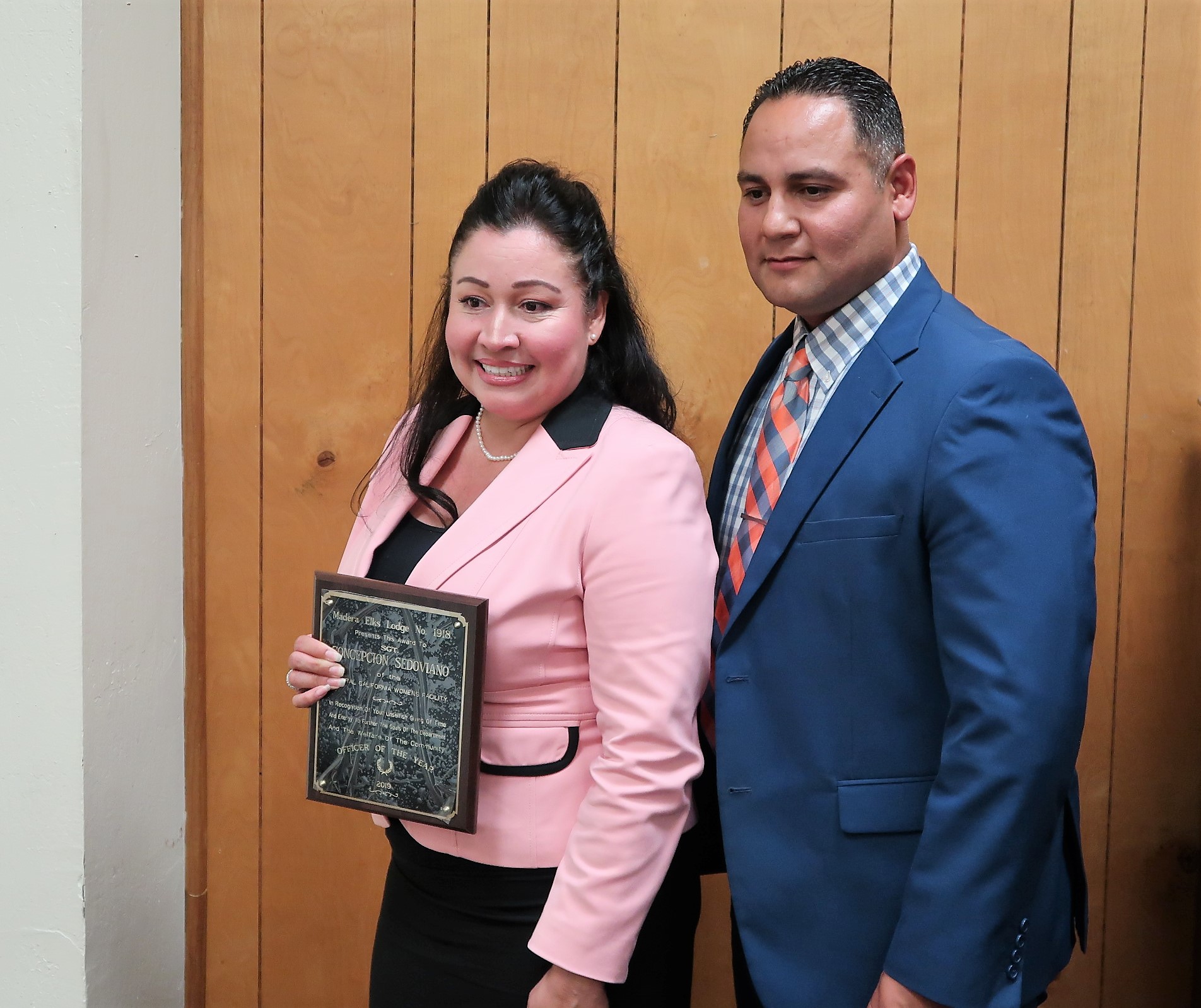 Woman in pink blazer and man in suit and tie pose for a photo. She is holding a plaque.