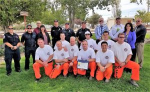 Inmates wearing orange and white sit on the ground while one holds a certificate of thanks. Standing around them are people in business attire and uniforms.