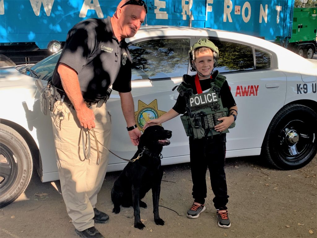 K9 officer with dog and boy wearing helmet and vest, all in front of a law enforcement vehicle.