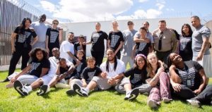 Inmates and kids sit in a yard surrounded by a fence with barbed wire.