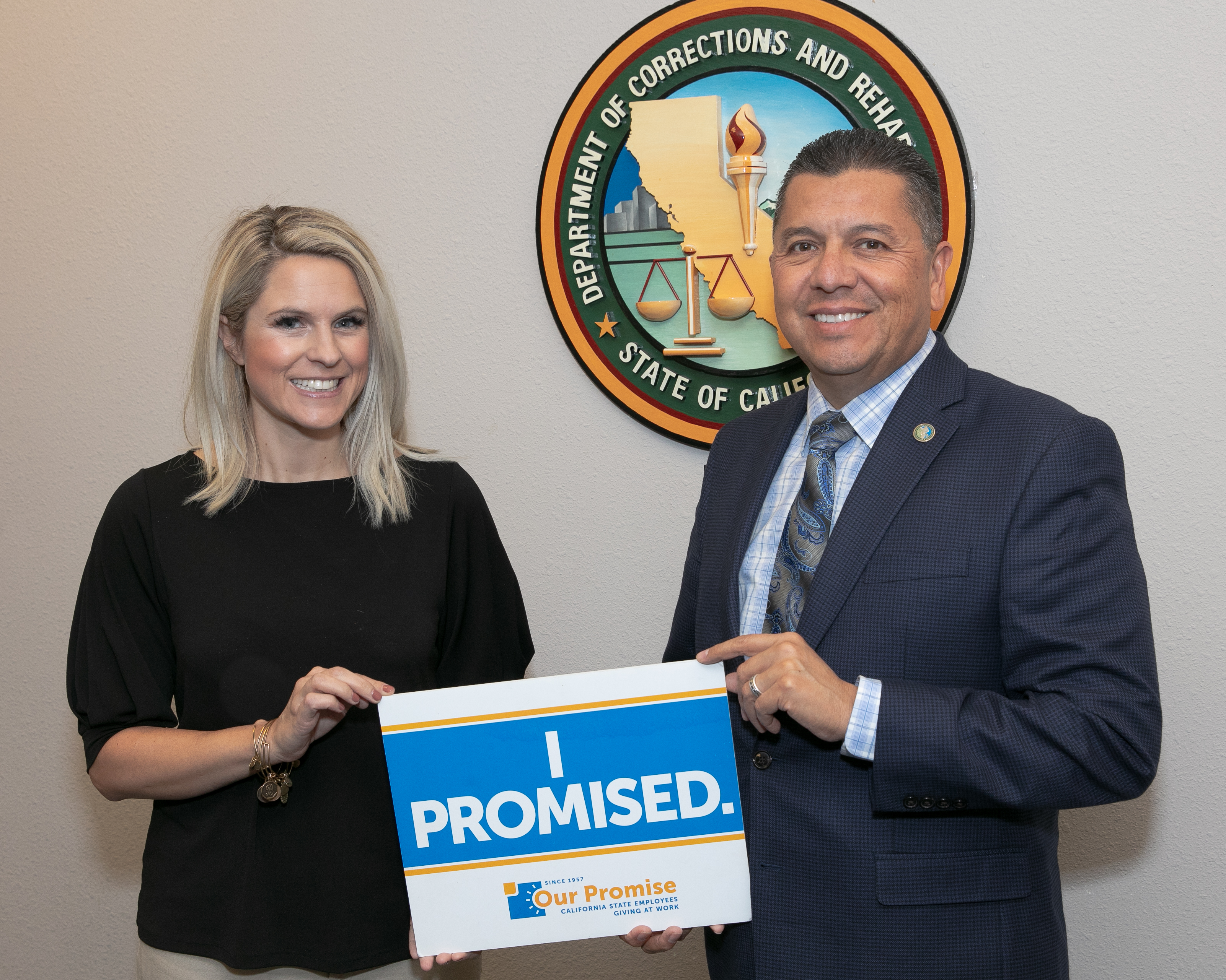 A man and woman face the camera while holding a sign that says "I Promised." Behind them is the CDCR logo.