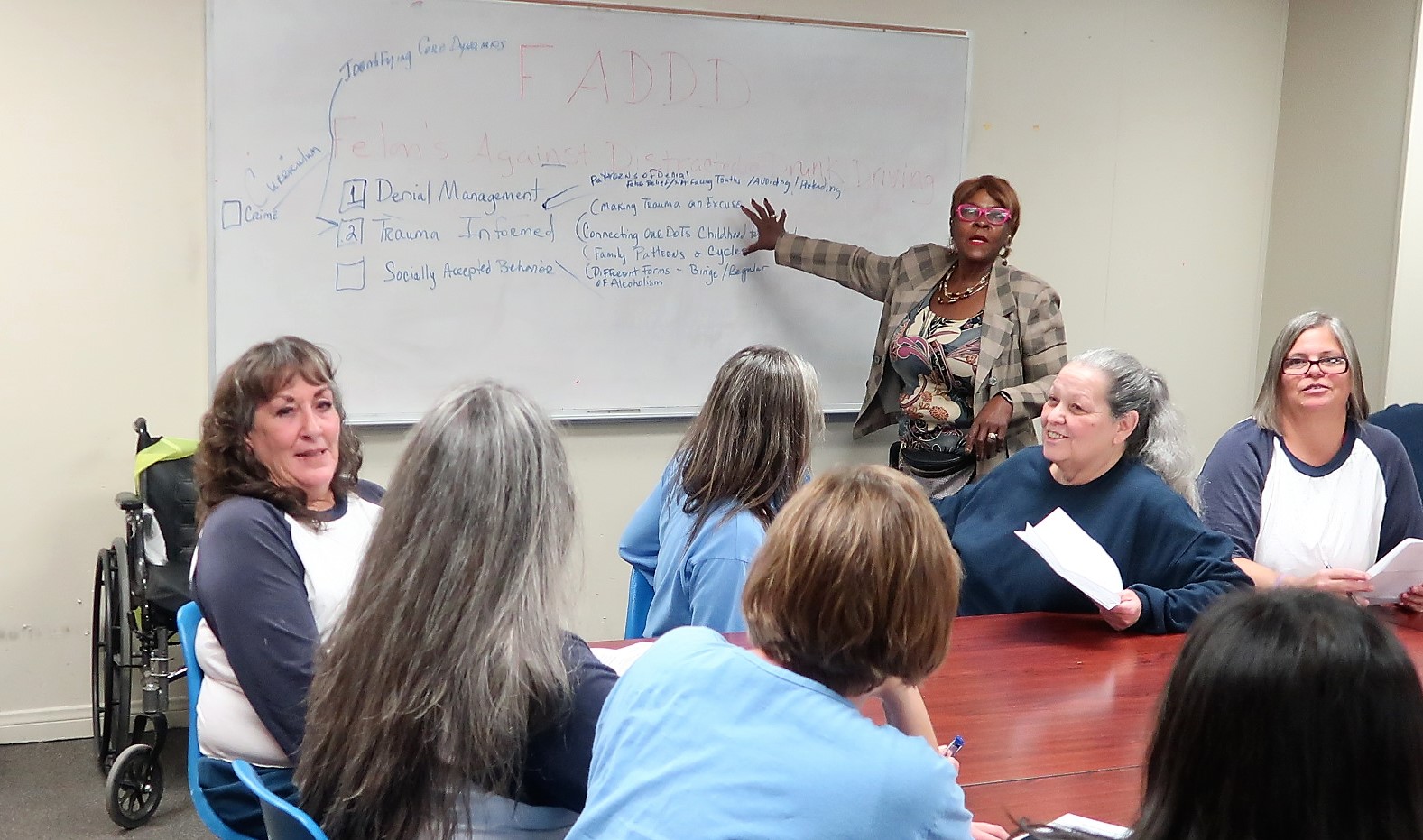 One woman points to a dry-erase board while others sit around a table.