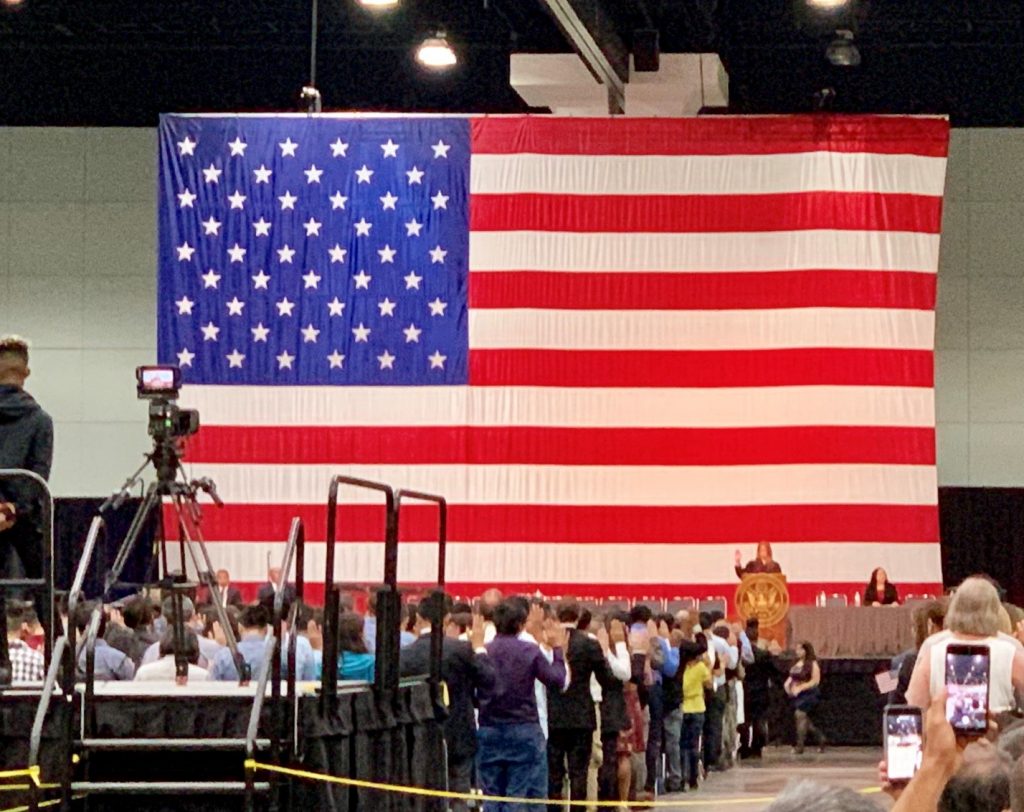 Huge American flag on stage with large crowd pledge oath using one hand raised 