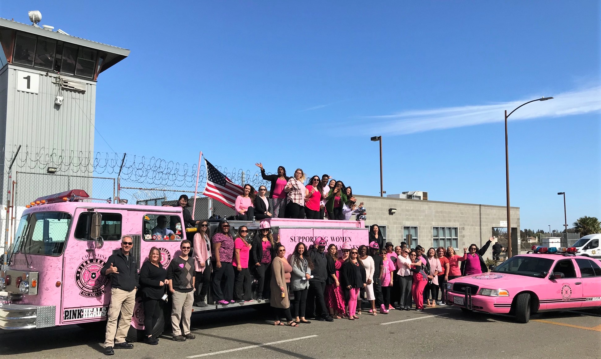 People wear pink and stand on top of a pink fire engine. A prison tower and fence can be seen in the background.