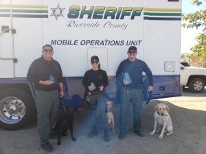 Three officers and their dogs stand in front of a Riverside County Sheriff mobile operations unit van.