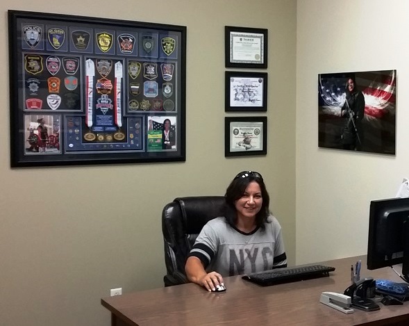 Woman sits at desk. On the walls are plaques and photographs.