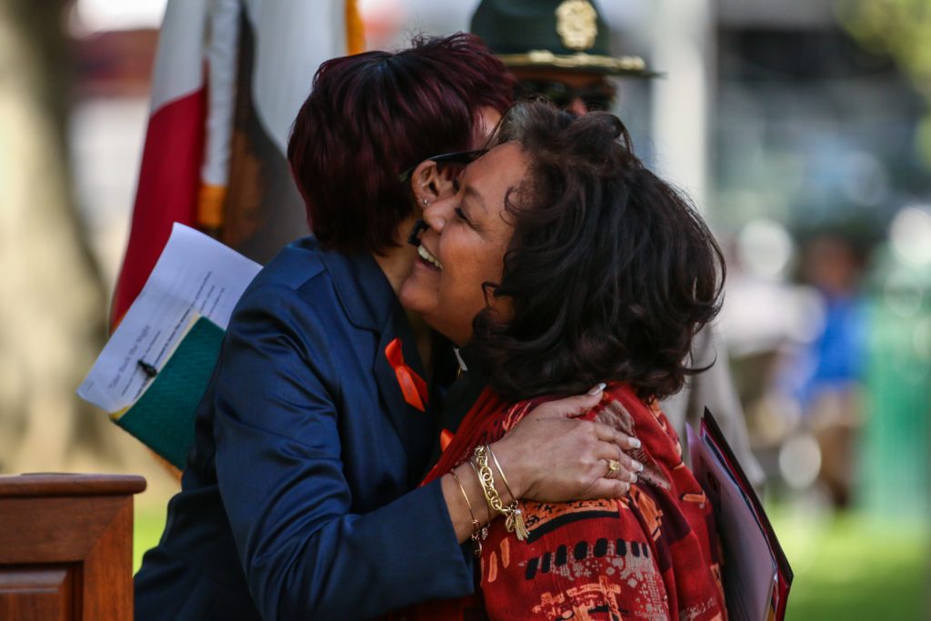 Two women embrace with a flag behind them.