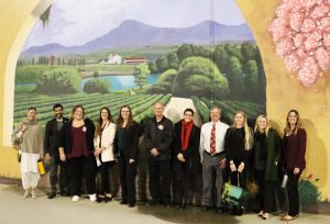 Men and women stand in front of a large mural depicting agriculture and a lake.