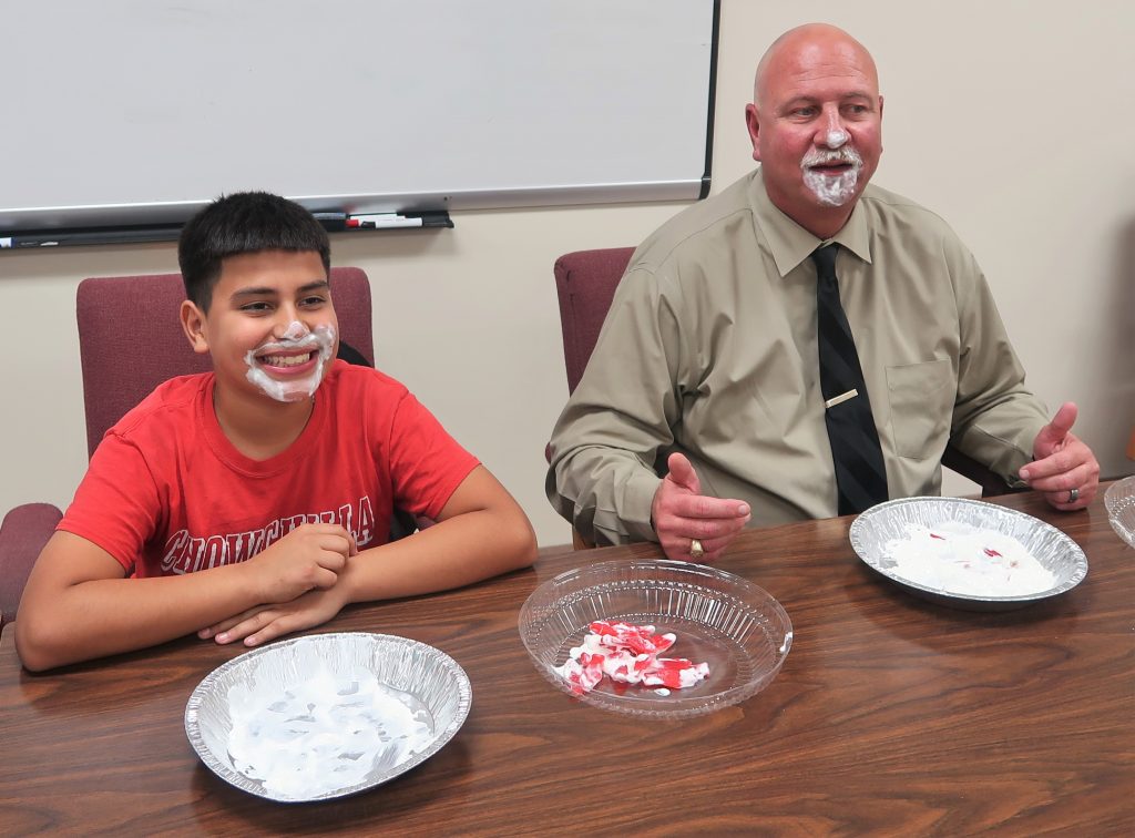 Prison executive and kid sit at table with whipped cream on their faces after a pie eating contest.
