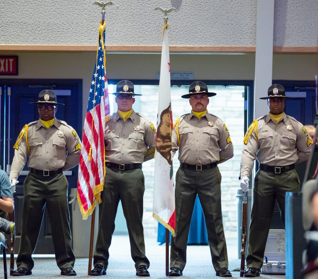 Four uniformed officers facing camera with one holding U.S. flag and another holding California flag.