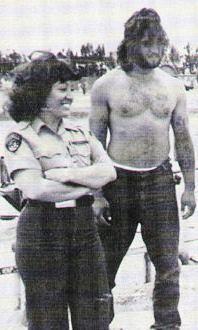 Female correctional officer stands in a prison yard next to a shirtless male inmate.
