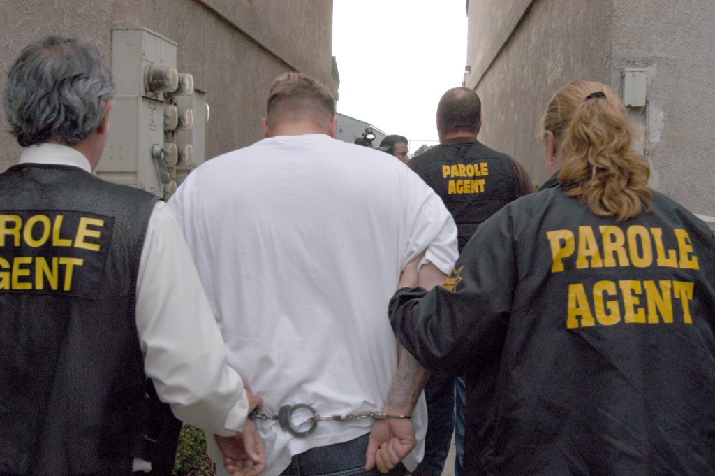 Men in women wear "parole agent" jackets while escorting a handcuffed man.