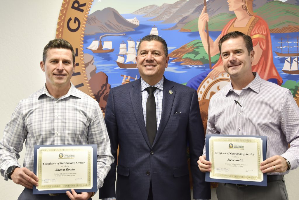 Two men hold certificates. They pose with a third man in front of the California State Seal.
