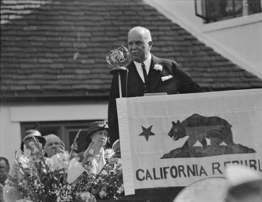 California governor James Rolph speaks at the microphone while standing behind a California flag.