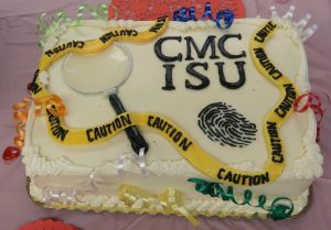 Fingerprint, caution tape and magnifying glass highlight a cake with the letters CMC ISU on it.