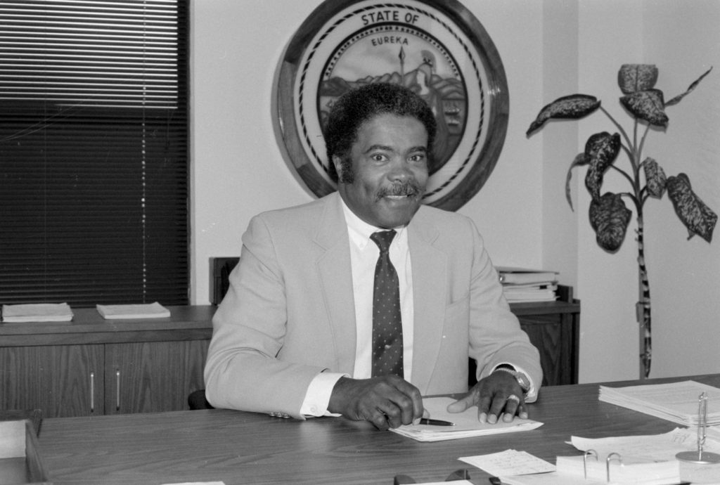 Man in tan suit and tie sits at a desk with the seal of California logo behind him.