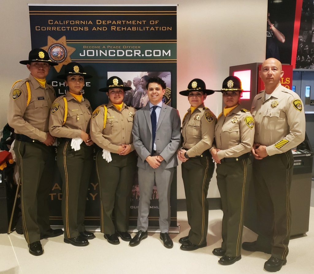Six correctional officers and one man in a suit stand in front of a poster that reads JoinCDCR.com.