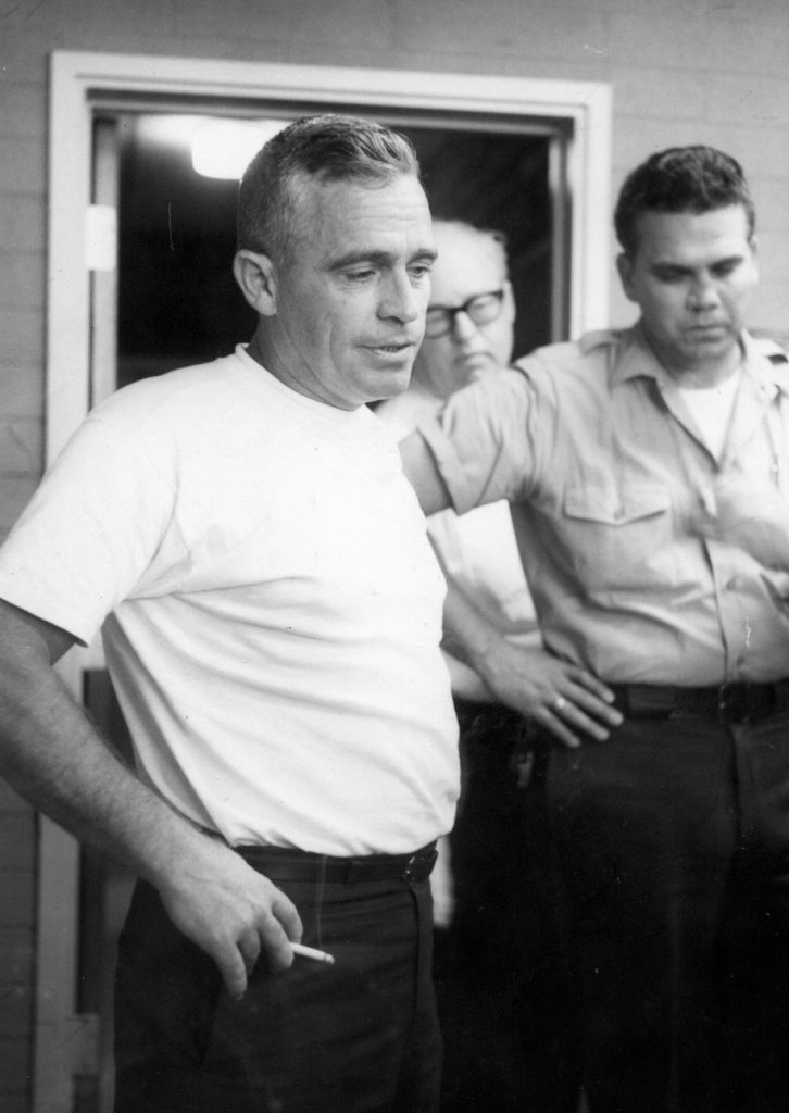 Man in t-shirt, holding a cigarette, and another man in uniform.