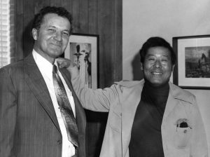 Two man stand in an office, smiling for the camera.
