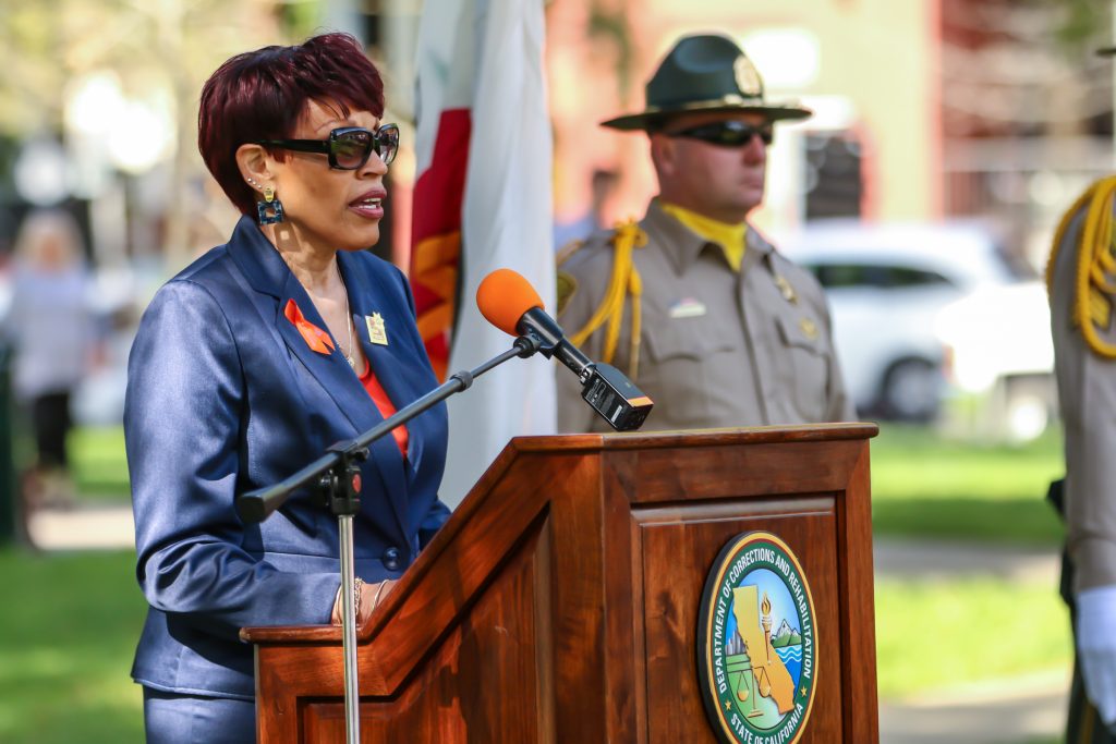 Woman speaks at lectern while CDCR honor guard officers stand in the background at a park.