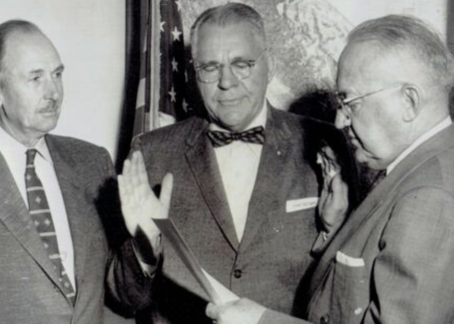 A man raises is right hand while standing in front of a US flag and map of California.